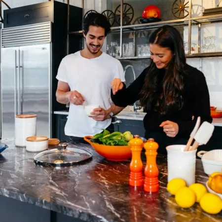 A smiling couple prepares a meal together in a modern kitchen with a black marble countertop, stainless steel appliances, and open shelving with glassware and cookware. Bright orange and yellow kitchen accessories, including a bowl of lemons and pepper grinders, add warmth to the scene.