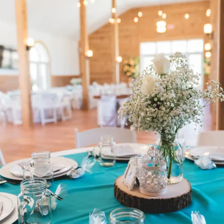 Rustic wedding reception table with a turquoise tablecloth, featuring a centerpiece of baby’s breath and white roses in a mason jar, placed on a wooden slab alongside a decorative lantern and table number.