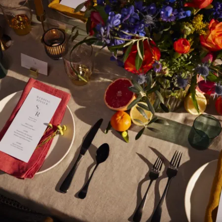 A beautifully set wedding dinner table featuring a menu with the initials “S/R” placed on a red cloth napkin on a white plate. The table is adorned with vibrant flowers in shades of blue, purple, orange, and yellow, accompanied by citrus fruits like oranges, lemons, and grapefruits as part of the centerpiece. Black cutlery is neatly arranged alongside green glass cups and lit candles, creating a warm and inviting atmosphere for the celebration.