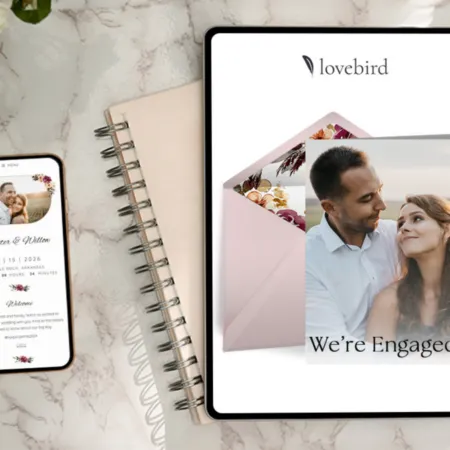 A digital wedding planning setup featuring a smartphone displaying a wedding website for "Hunter & Wilton" with a countdown to the event, a tablet with an open engagement announcement displaying a couple, and a pink envelope with floral details on a marble surface.
