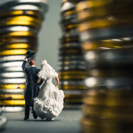 A miniature bride and groom figurine stand together, surrounded by towering stacks of gold and silver coins, symbolizing the financial considerations and expenses associated with weddings. The groom lifts his top hat in a celebratory pose, while the bride holds her dress delicately, walking arm-in-arm toward their future.