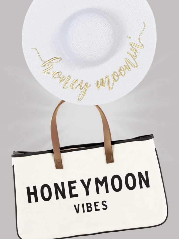 White wide-brimmed hat with "Honeymoonin'" written in gold and a canvas tote bag with "Honeymoon Vibes" written in bold black letters.