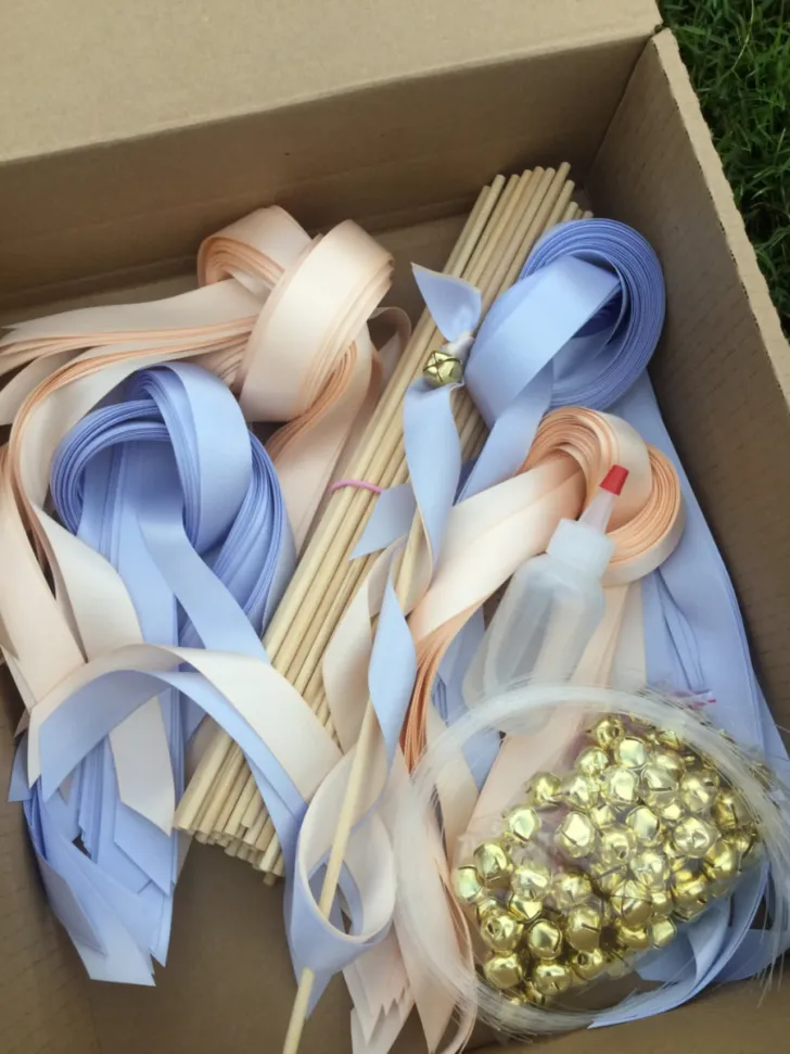 A box filled with wedding ribbon wands, showcasing soft blue and pink ribbons attached to light wooden sticks, complemented by golden bell embellishments for a cheerful sound during the celebration.